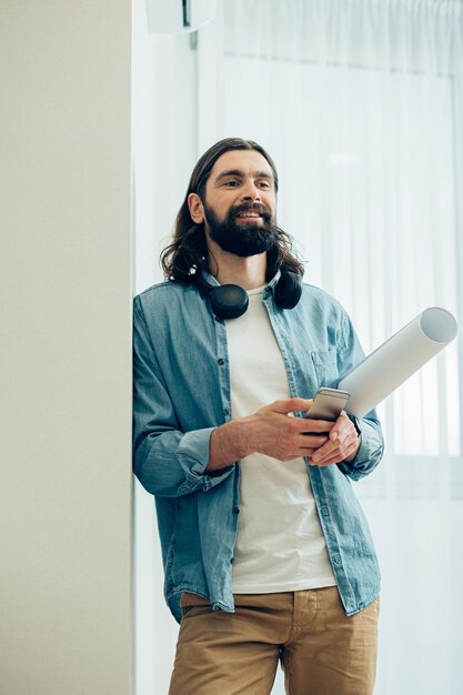 Mirthful young man in casual clothes standing indoors with a roll of drawing paper and a smartphone