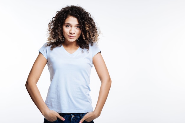 Mischievous tomboy. Beautiful curly young woman in a white t-shirt holding her hands in the pockets of her jeans while posing on grey background