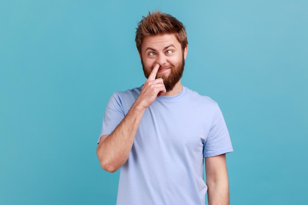 Misconduct, bad manners. Portrait of crazy bearded man picking nose and sticking out tongue with dumb comical expression, fooling around. Indoor studio shot isolated on blue background.