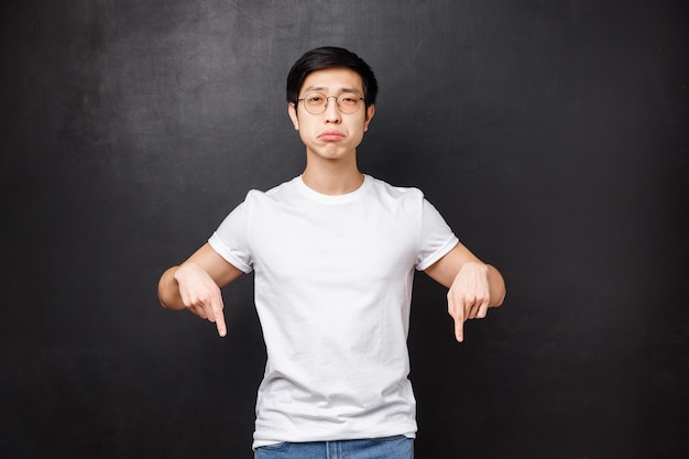 Miserable and gloomy upset cute asian guy sobbing look at camera with regret and sadness, pointing fingers down at something broken or upsetting, feel uneasy, 