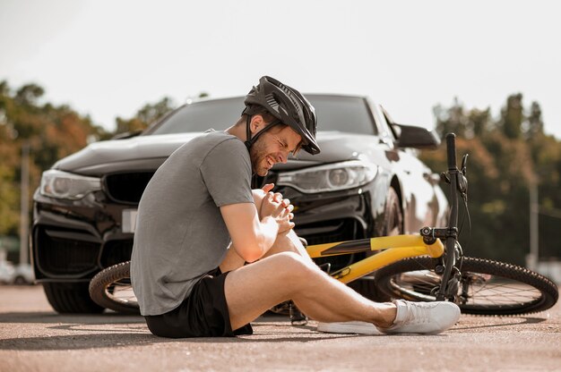 Photo misfortune. unhappy guy in helmet sitting with closed eyes holding his knee near bicycle on road in front of car
