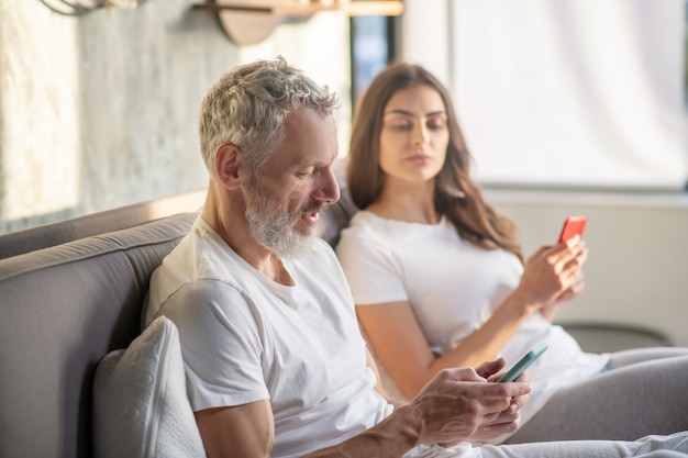 Mistrust. Adult bearded husband staring at smartphone and peeping distrustful wife sitting side by side on bed