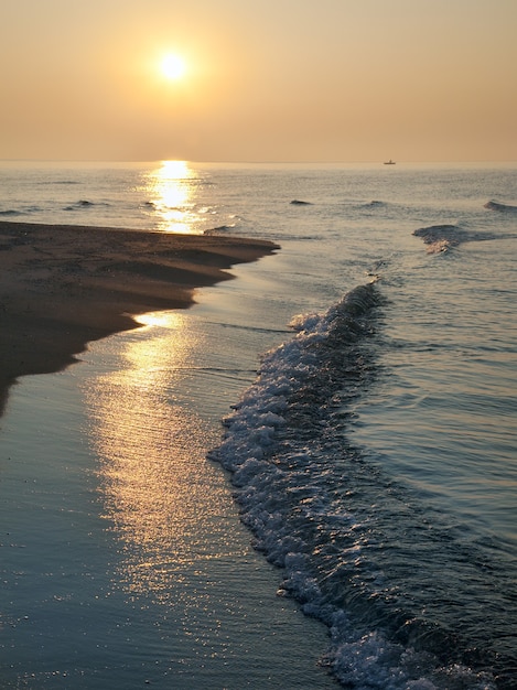 Misty daybreak sea sandy shore with sunlight path and fishing boat distant silhouette