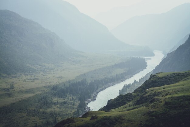 Misty mountain landscape with wide mountain river. Dark green gloomy scenery with confluence of two mountain river in mist. Dark atmospheric view to confluence of great rivers in rainy weather.