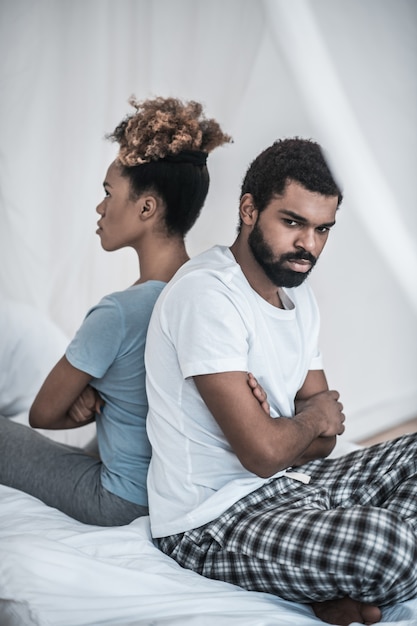 Photo misunderstanding. young adult dark-skinned angry man and woman in pajamas sitting with backs to each other on bed at home