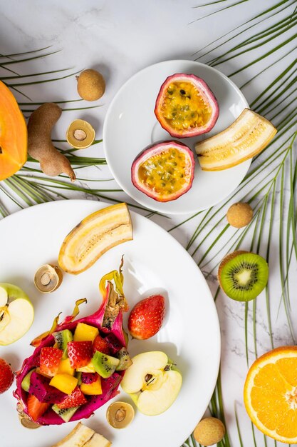 Mix of different tropical fresh ripe fruits on white plate and on table with green palm leaves. Exotic breakfast, delicious snack. Summer, vacation and healthy eating concept. Top view, vertical shot.