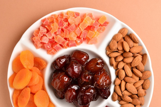 Mix of dried fruits and nuts on a white plate. Apricot, almond, raisin, dates fruit. On a brown background. Space for text or design.