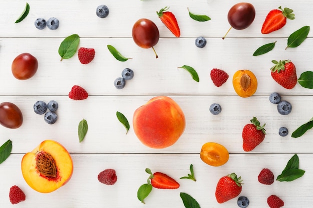 Mix of organic berries background. Pattern of fresh strawberries, plums, raspberries, peaches, blueberries and mint leaves on white wooden table, top view