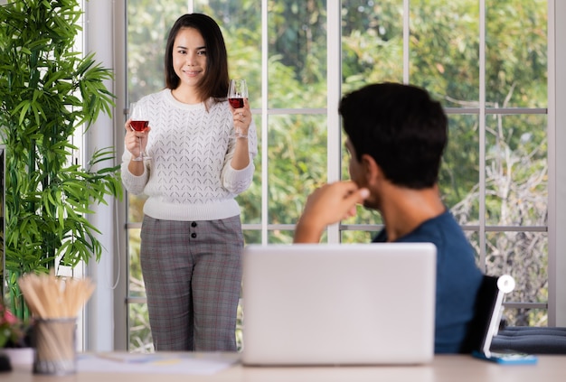 Mix race family lovers, Caucasian husband and Asian wife, man working at desk with laptop computer and woman taking couple of red wine glass to celebrate and relaxing. Concept of love and care at home