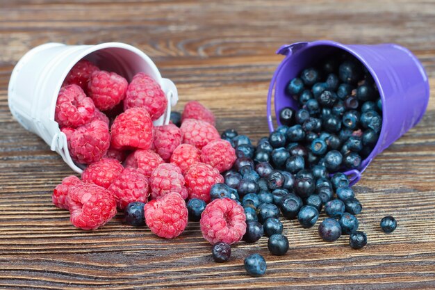 Mix of raspberries and blueberries in a small white and blue buckets on brown wooden background.