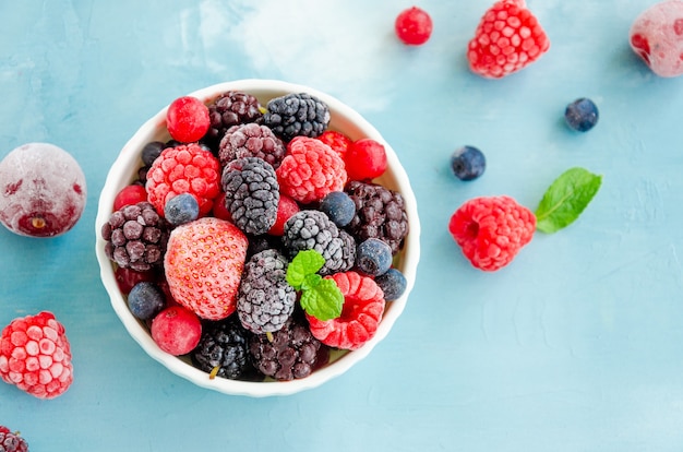 Mix of summer frozen berries in a white bowl on a blue concrete background. Copy space.