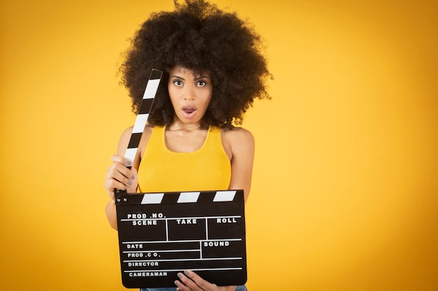 Mixed afro american young woman, excited, casual wear, keeping mouth open hold classic black movie clapperboard isolated on orange background.