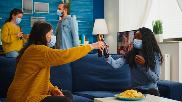 Mixed race black woman taking off protection masks clinking bottles of beer, drinking and eating snacks spending free time in living room respecting social distance. Diverse people enjoying party in o