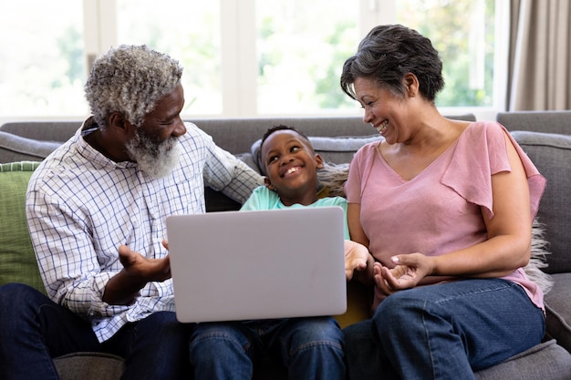 혼혈 소년과 그의 조부모는 집에서 함께 시간을 즐기고, 소파에 앉아, 노트북을 사용하고, 서로를 바라보며 웃고 있습니다.