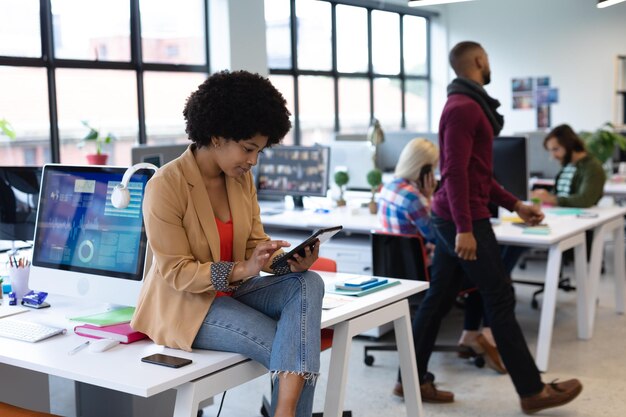 Mixed race businesswoman working in creative office. woman sitting on desk and using tablet computer, with colleagues in the background. business people and work colleagues at a busy creative office.