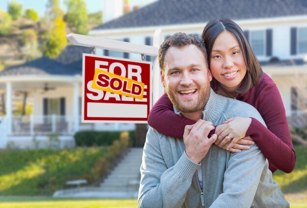 Mixed Race Caucasian and Chinese Couple In Front of Sold For Sale Real Estate Sign and House