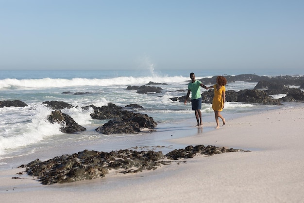 A mixed race couple enjoying free time on beach on a sunny day together, holding hands, having fun and laughing with sun shining on their faces. Relaxing summer vacation.