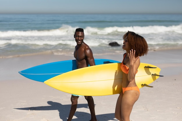 A mixed race couple enjoying free time on beach on a sunny day together, surfing, carrying their surfboards, having fun with sun shining on their faces.