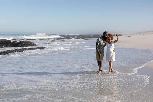 A mixed race couple enjoying free time on beach on a sunny day together, walking, taking photos with a smartphone and holding each other with sun shining on their faces.