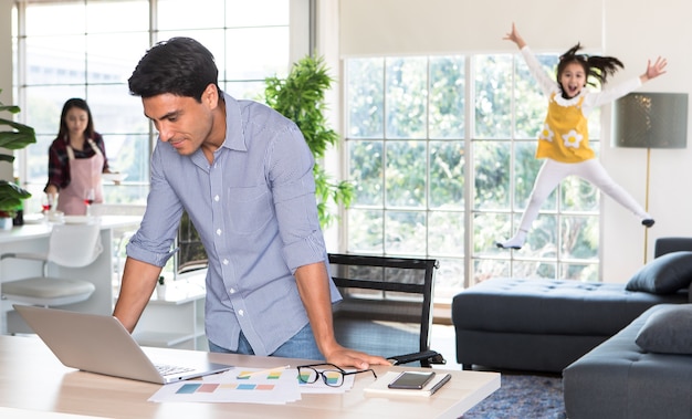 Mixed race family staying together, Caucasian dad working at home at desk while mother prepare food and drink in kitchen and little daughter play and jumping on sofa in blur, idea for warm family.