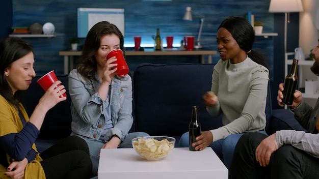 Mixed race friends cheering bottles of beer enjoying time spend together while relaxing on sofa. Group of multi ethnic people hanging out, socializing during wekeend party late at night