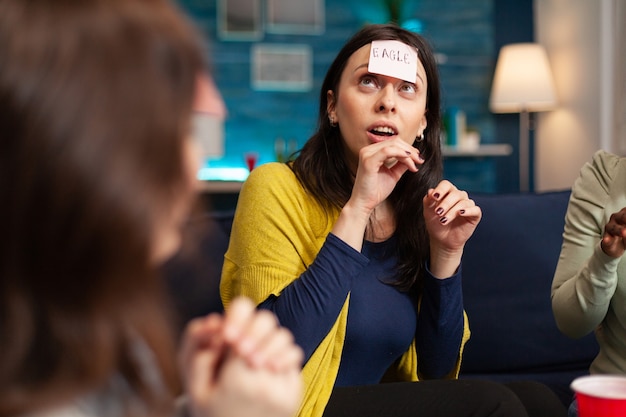 Mixed race friends with sticky notes attached on forehead playing guess who game during home party. Multi-ethnic people having fun, laughing together while sitting on sofa in living room late at night