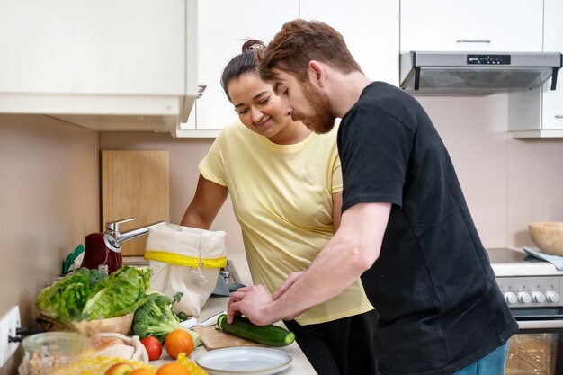 Mixed-race happy young couple preparing vegan food. Attractive caucasian man and mixed-race woman preparing vegan meal at kitchen. Vegan natural food cooking. Comfort cooking. Real life concept