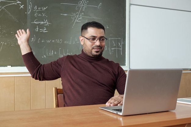 机のそばに座ってノートパソコンのディスプレイを見ながら黒板の方程式の解を指しているカジュアルウェアの混血男性教師