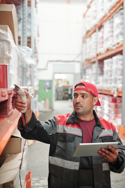 Mixed-race male worker of warehouse in uniform scanning qr codes on boxes containing goods while standing in aisle by huge shelf