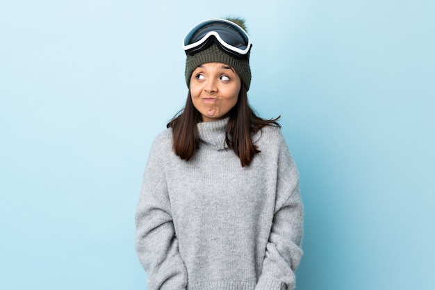 Mixed race skier girl with snowboarding glasses over blue wall having doubts while looking up.