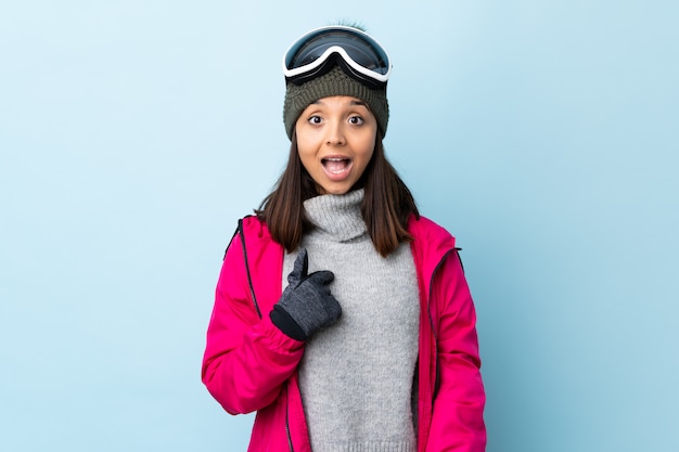 驚いた表情で青い壁の上のスノーボードメガネと混血スキーヤー少女