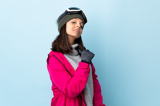 Mixed race skier girl with snowboarding glasses over isolated blue background proud and self-satisfied