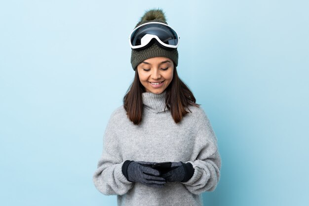 Photo mixed race skier girl with snowboarding glasses over isolated blue background sending a message with the mobile