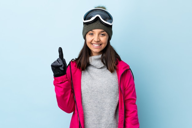 Mixed race skier girl with snowboarding glasses over isolated blue background