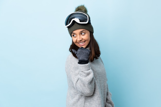 Mixed race skier girl with snowboarding glasses over isolated blue pointing to the side to present a product.