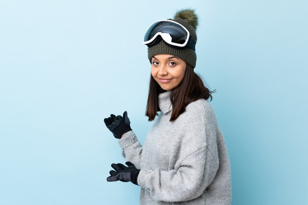 Mixed race skier girl with snowboarding glasses over isolated blue wall extending hands to the side for inviting to come