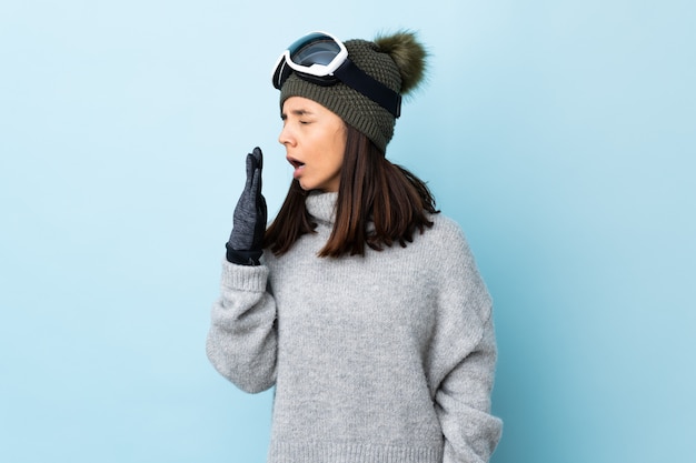 Mixed race skier woman with snowboarding glasses over isolated blue space yawning and covering wide open mouth with hand