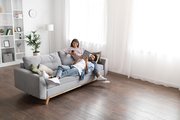 Mixed race spouses resting on sofa at home using gadgets