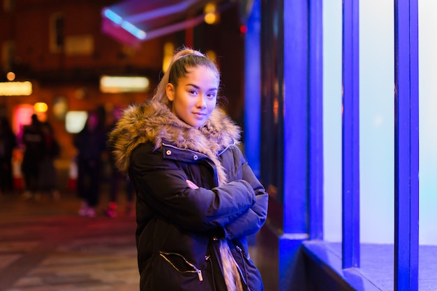 Mixed race teenage with fur coat standing next to window with blue light