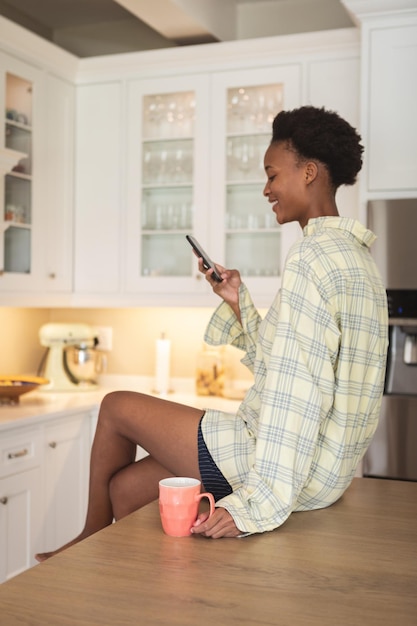 Mixed race woman spending time at home, sitting in her kitchen and using a smartphone. self isolating and social distancing in quarantine lockdown during coronavirus covid 19 epidemic