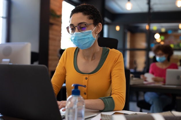 Photo mixed race woman wearing face mask using laptop at desk in casual office. sanitizing gel on her desk. hygiene in workplace during coronavirus covid 19 pandemic.