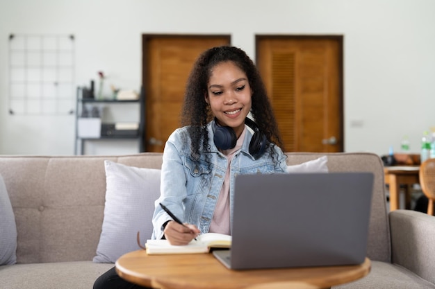 Mixed race woman wearing headphones watching webinar write\
notes study online with online teacher african female student\
learning language computer course on laptop listen translate\
lecture