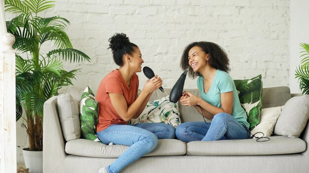 Mixed race young funny girls dance singing with hairdryer and comb sitting on sofa