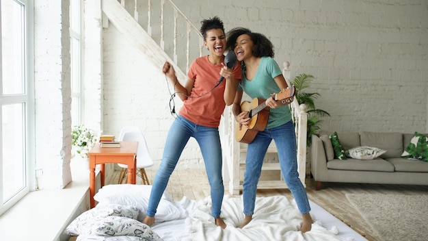 Mixed race young funny girls dance singing with hairdryer and playing acoustic guitar on a bed