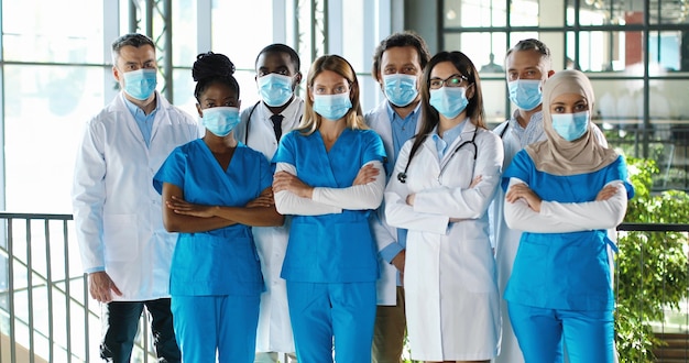 Photo mixed-races busy team of male and female doctors posing to camera and crossing hands in hospital. international group of medics in medical masks. protected multi ethnic physicians and nurses in clinic