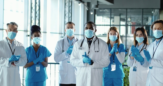 mixed-races team of male and female doctors applauding in hospital. international group of medics in medical masks. protected workers. multi ethnic physicians and nurses in uniforms in clinic applause
