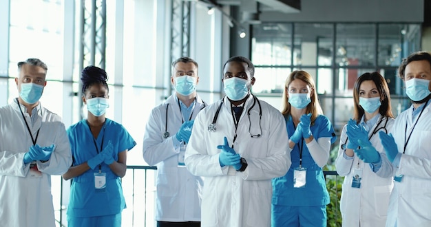 Mixed-races team of male and female doctors applauding in hospital. International group of medics in medical masks. Protected workers. Multi ethnic physicians and nurses in uniforms in clinic Applause