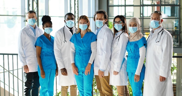 Mixed-races team of specialists, males and females doctors in hospital. International group of medics in medical masks. Protected workers. Multi ethnic physicians and nurses in uniforms in clinic.