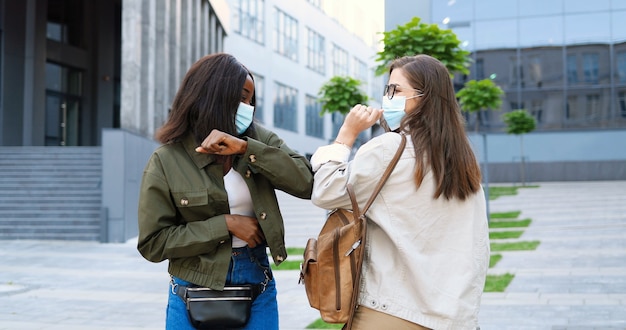 Mixed-races young joyful females friends in medical masks meeting at street and greeting with elbows. Multi ethnic happy girls talking and chatting cheerfully African American and Caucasian students.