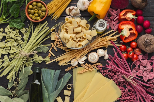 Mixed raw colorful italian pasta background. Handmade macaroni with various vegetables and spices on dark wood, top view, copy space